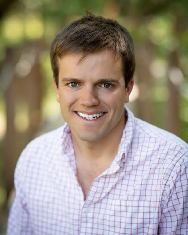 Headshot of a smiling man in a pink checkered button-up shirt outdoors