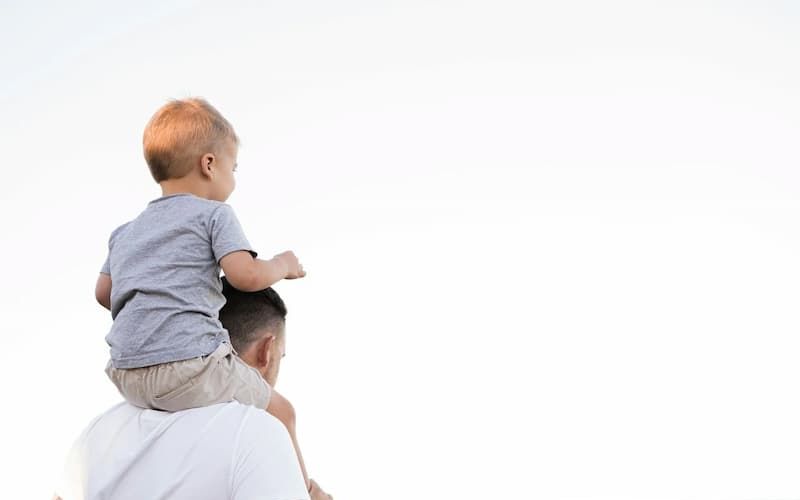 Toddler riding on a father's shoulders against a bright sky