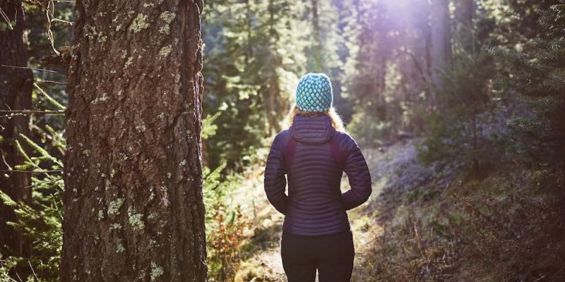 Woman in a knit beanie and puffer jacket hiking a sunlit forest trail
