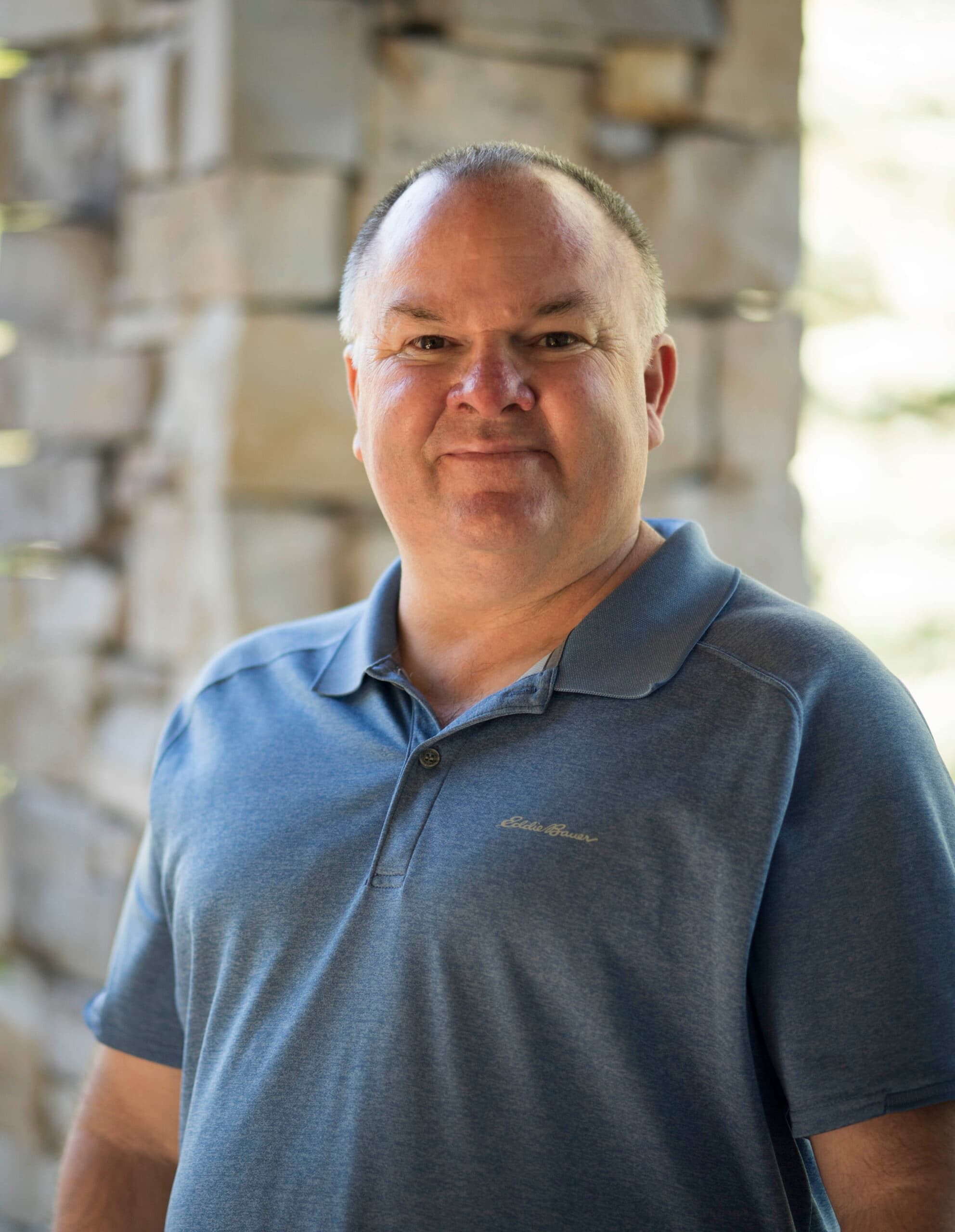 Smiling man in a blue polo shirt standing in front of a stone wall