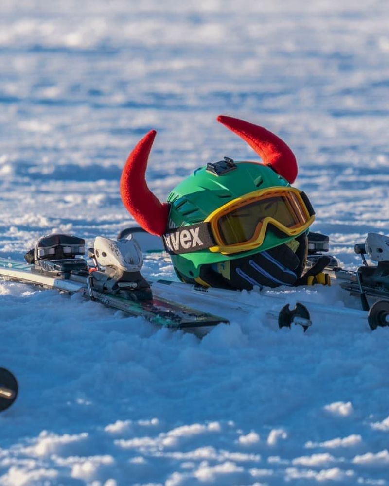 Green ski helmet with red horns and goggles resting on skis in snow