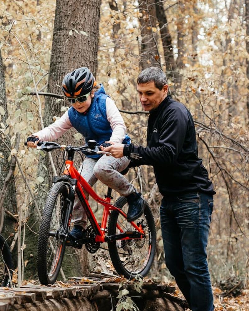 Father guiding a child riding a red mountain bike over a log on a forest trail