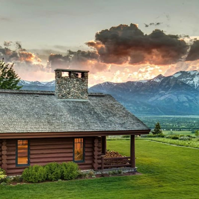 Log home with stone chimney at sunset overlooking distant snow-capped mountains