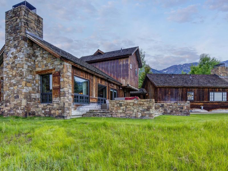 Stone-and-timber mountain home with grassy yard and distant peaks at dusk