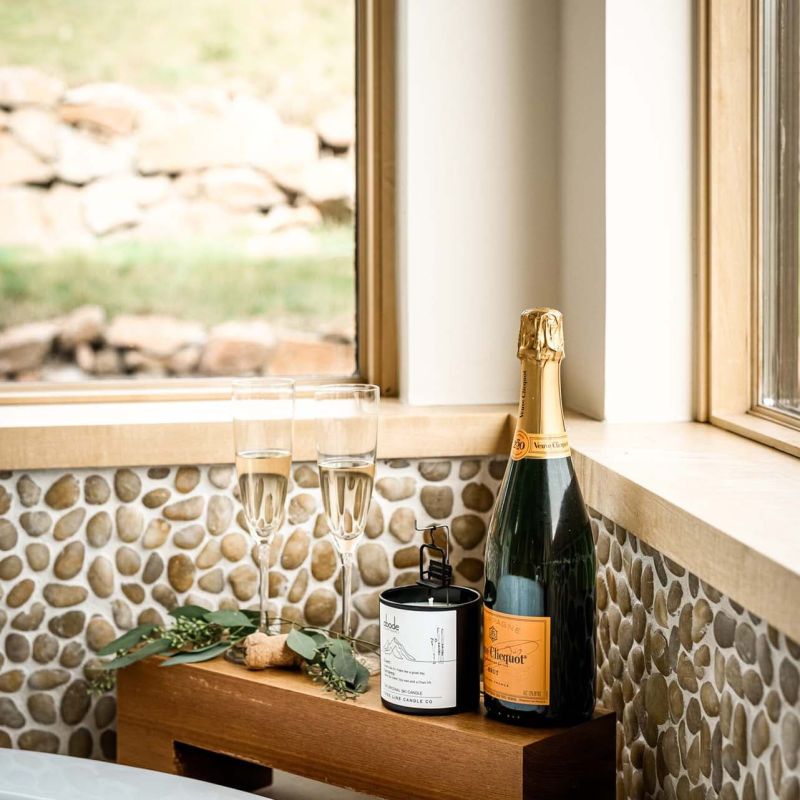 Champagne bottle and two flutes on a wooden tray beside a soaking tub with pebble-tile surround