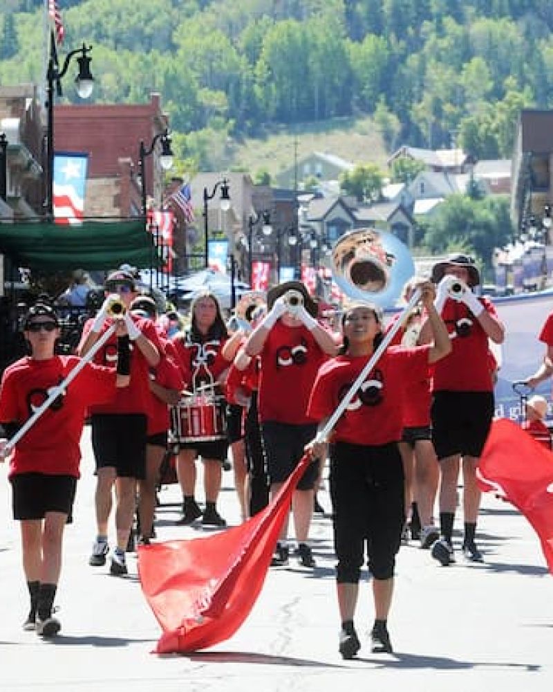 Marching band with red flags and brass instruments parading down Park City Main Street
