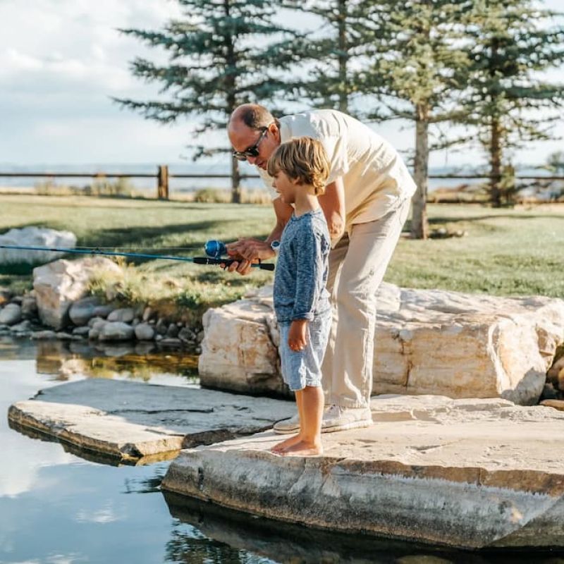 Grandfather and young child fishing from a stone ledge by a pond