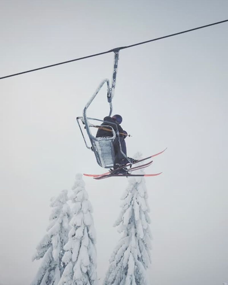 Skier riding a chairlift above snow-laden pine trees in foggy conditions