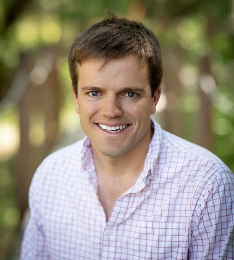 Headshot of a smiling man in a pink checkered button-up shirt outdoors