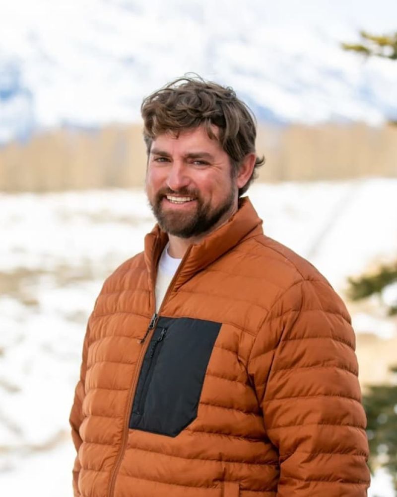 Bearded man in orange puffer jacket smiling outdoors with snowy mountains behind
