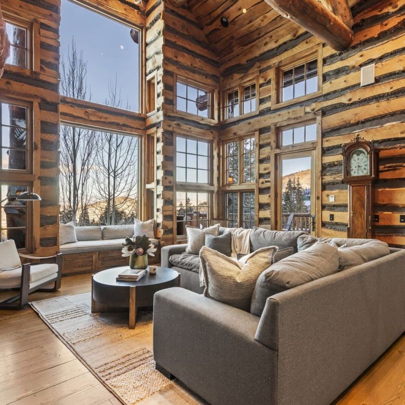 Log cabin great room with two-story windows facing snowy mountains
