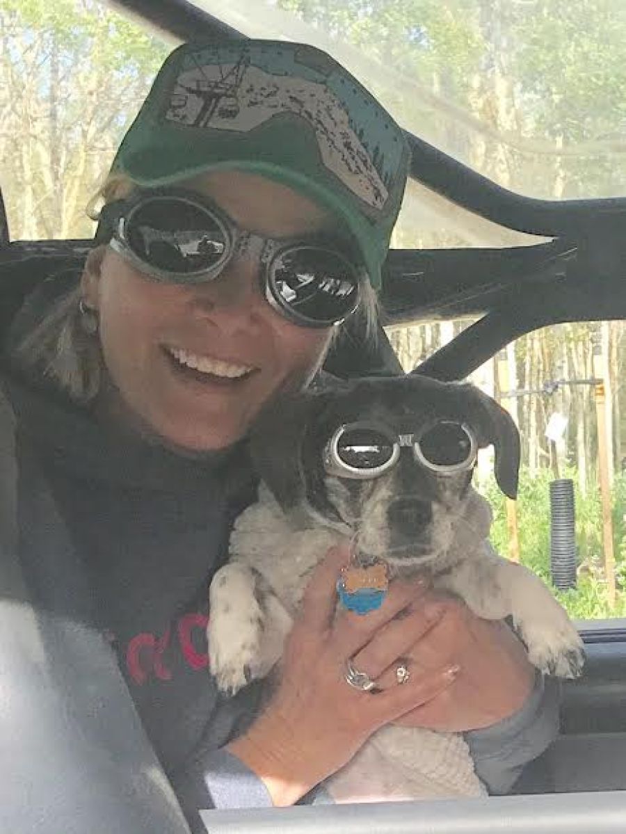 Smiling woman in sunglasses and ball cap holding a small dog wearing goggles