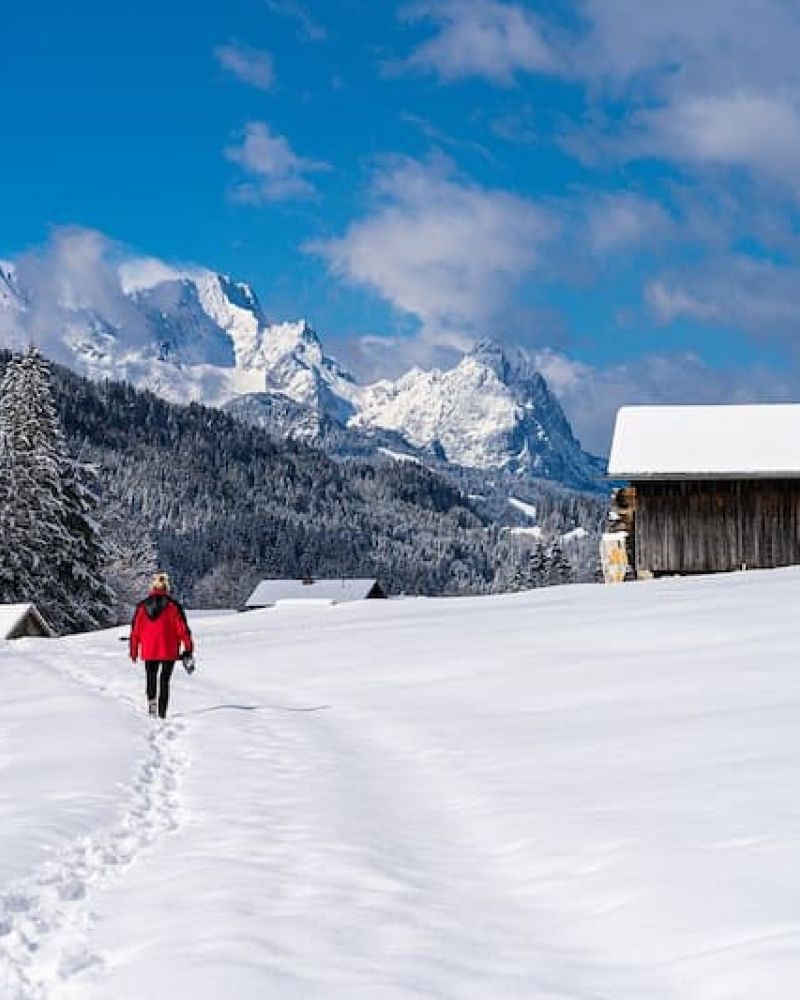 Hiker in red walking a snow-packed trail toward jagged alpine peaks