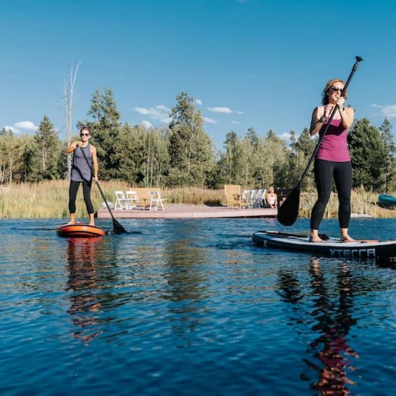 Two paddleboarders on calm water near a wooded shoreline and dock