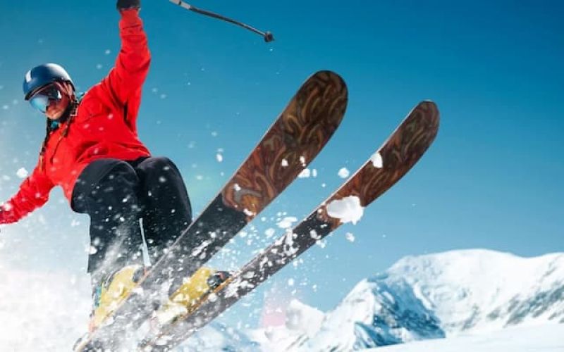 Skier in a red jacket catching air against snowy peaks and blue sky