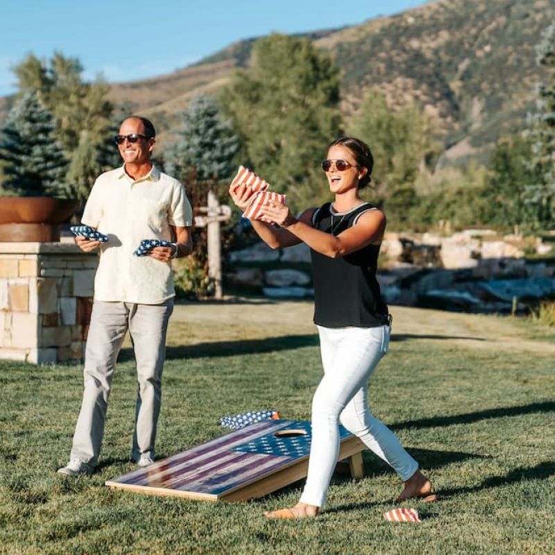 Two people playing cornhole on a grass lawn with mountain backdrop