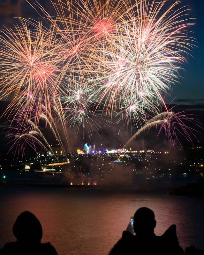 Silhouetted couple watching colorful fireworks burst over a harbor at dusk