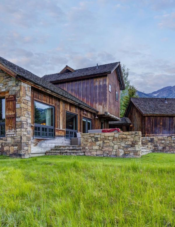 Stone-and-timber mountain home with grassy yard and distant peaks at dusk