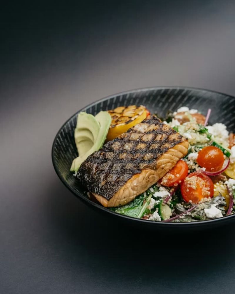 Grilled salmon fillet on a quinoa salad with cherry tomatoes, avocado, and feta in a black bowl