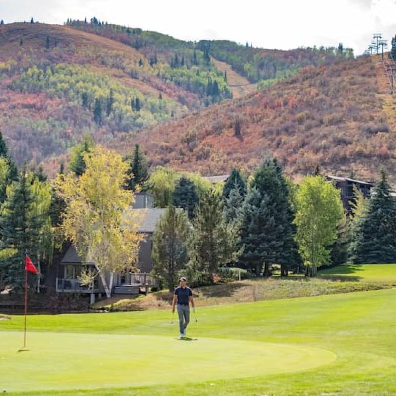 Golfer walking a putting green with autumn ski-run mountains behind