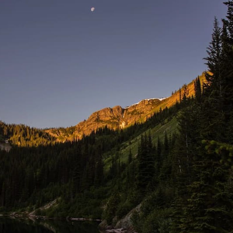 Forested mountain ridge glowing at sunset above a calm alpine lake under a moonlit sky