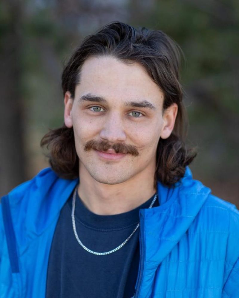 Headshot of a young man with a mustache wearing a blue jacket
