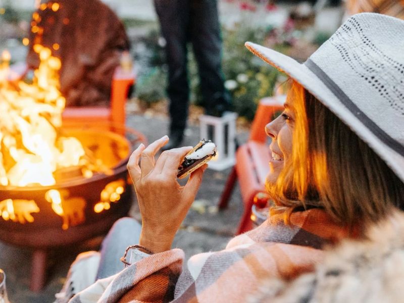 Woman in a wide-brim hat holding a s'more beside a flickering fire pit