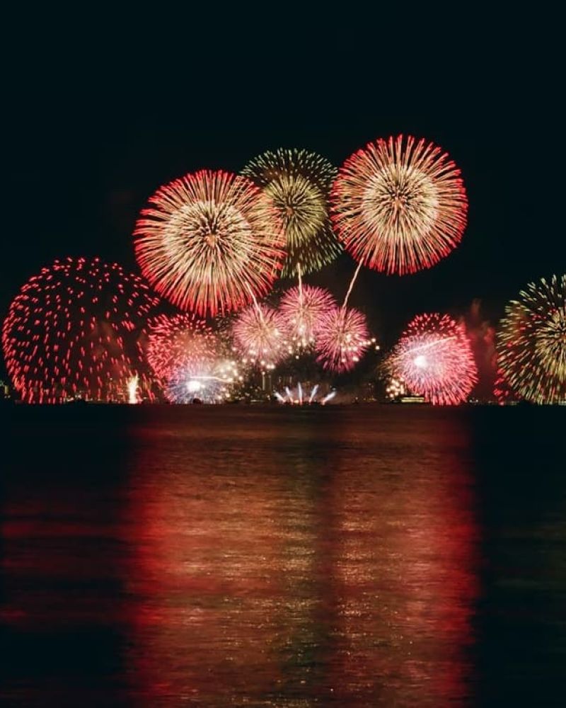 Large fireworks display reflected on dark water beside a city skyline