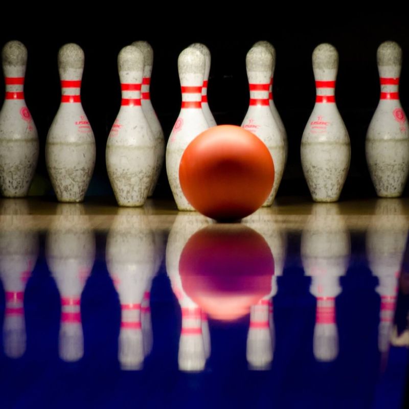 Bowling pins reflected on a polished lane with an orange ball mid-frame