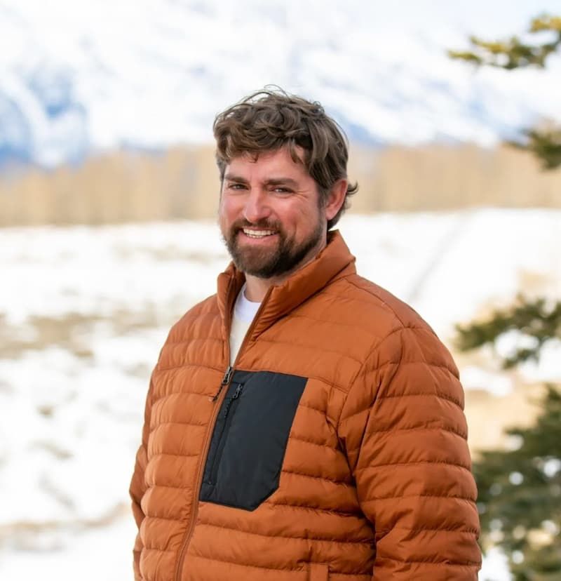 Bearded man in orange puffer jacket smiling outdoors with snowy mountains behind