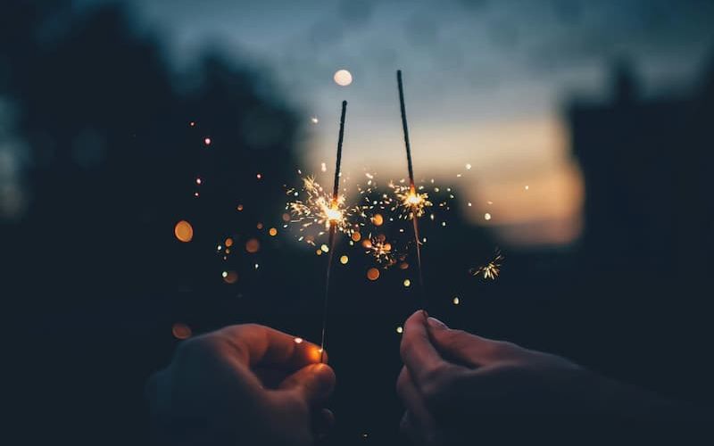 Two hands holding lit sparklers at dusk with bokeh lights behind