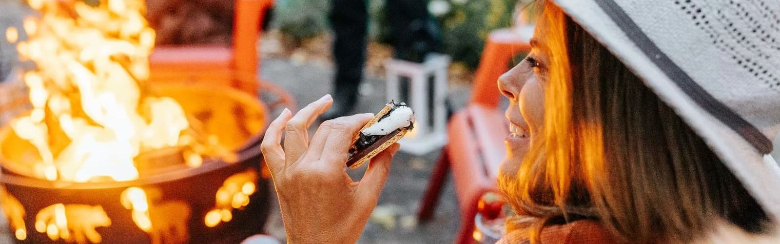 Woman in a wide-brim hat holding a s'more beside a flickering fire pit