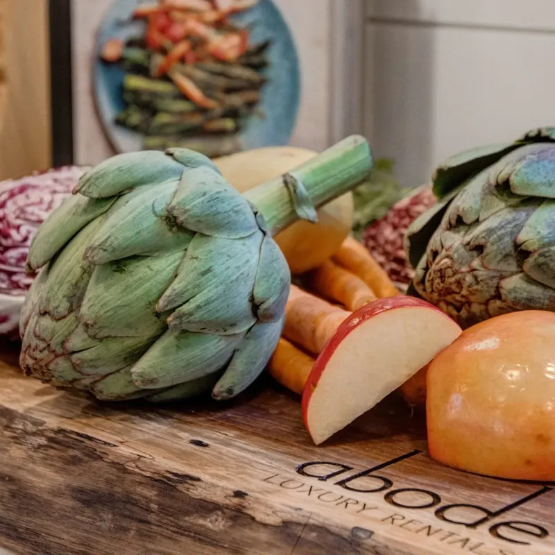 Fresh artichokes, apple, and carrots on a wooden cutting board with abode logo