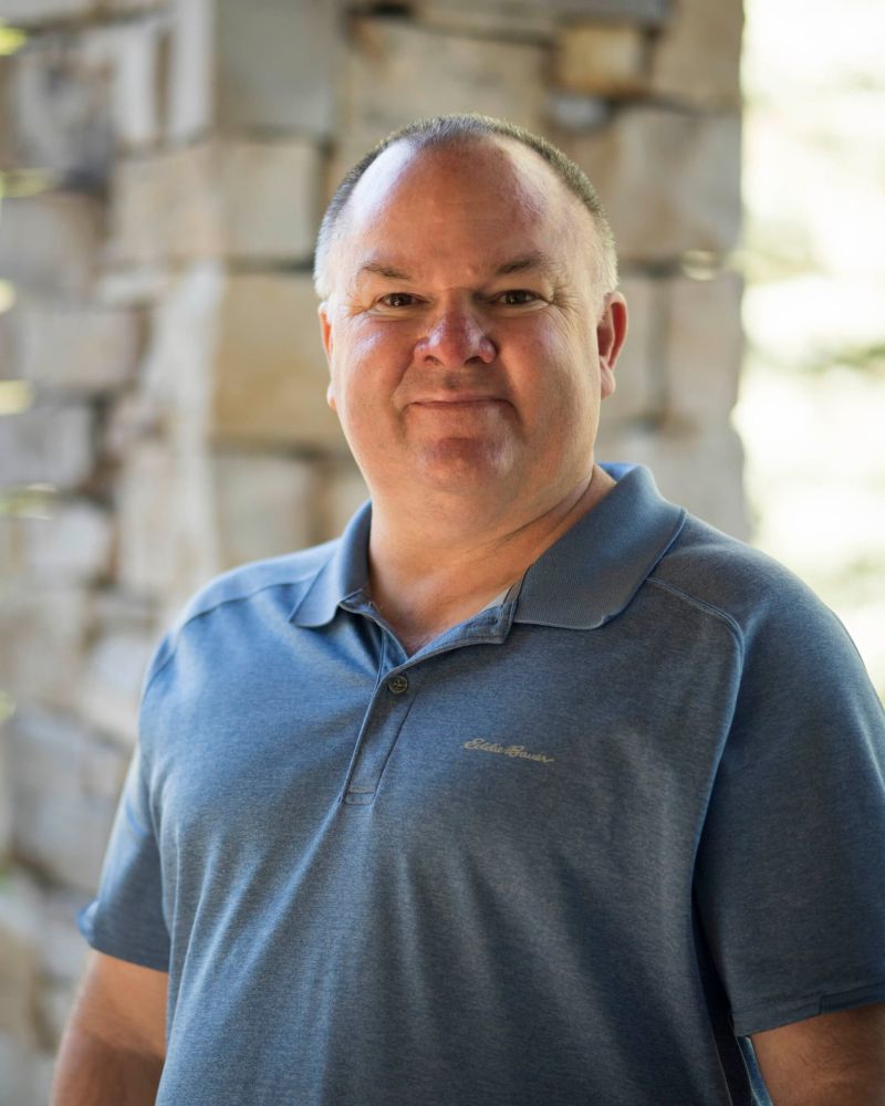 Smiling man in a blue polo shirt standing in front of a stone wall