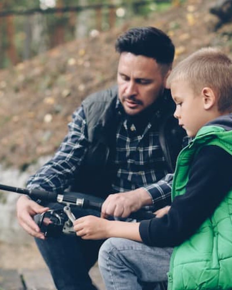 Father showing a young boy how to use a fishing reel by a wooded bank