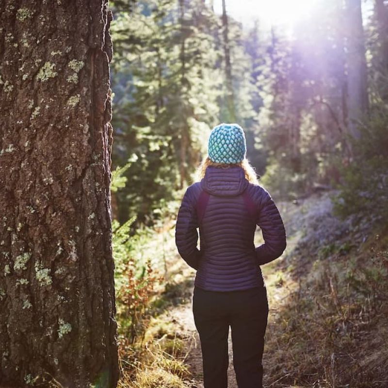 Woman in a knit beanie and puffer jacket hiking a sunlit forest trail
