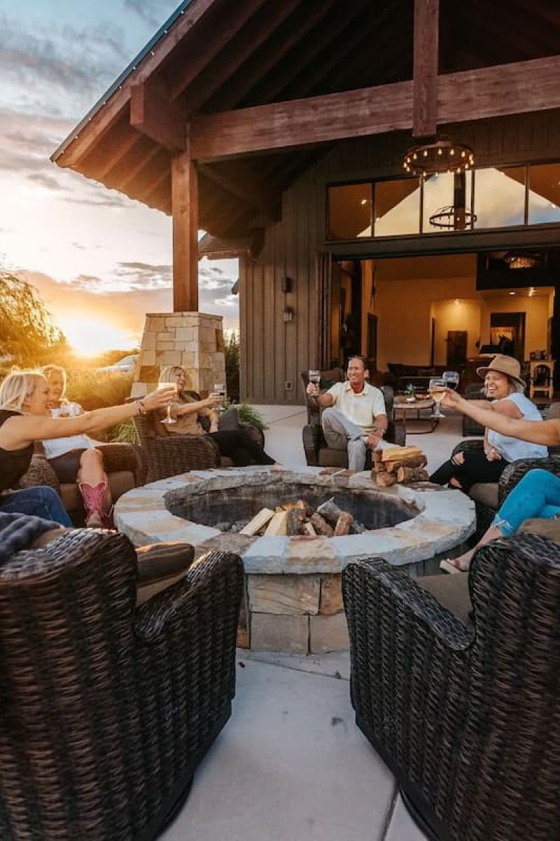 Group of friends toasting wine around an outdoor stone fire pit at sunset