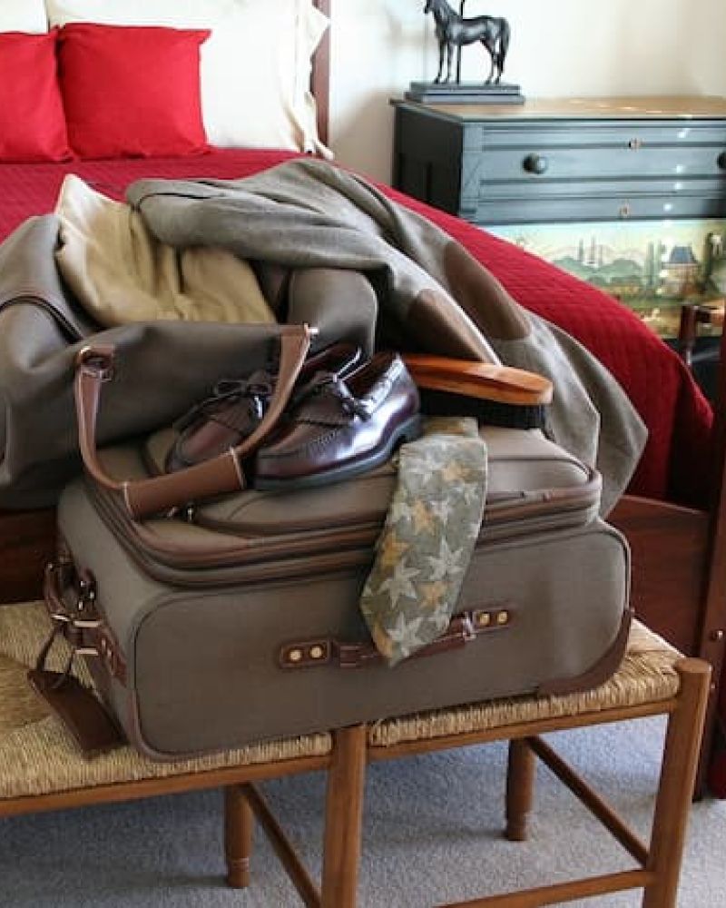 Packed travel suitcase with shoes and tie on a wooden bench beside a bed