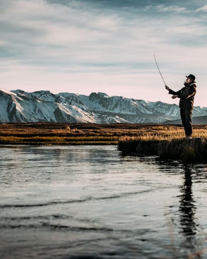 Fly fisherman casting a line in a meadow stream below snow-capped peaks