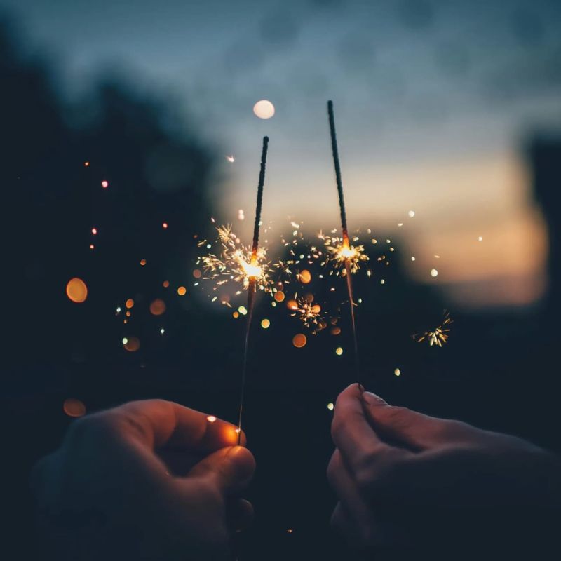 Two hands holding lit sparklers against a dusk sky