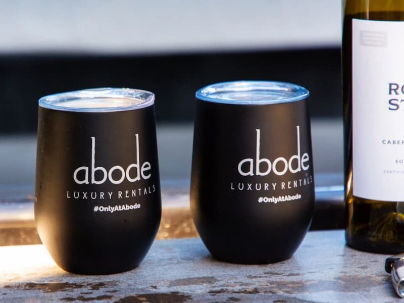 Two black Abode Luxury Rentals tumblers beside a bottle of wine