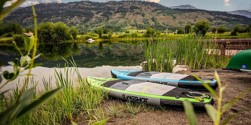 Two Slingshot paddleboards on a grassy lakeshore with mountains in the background