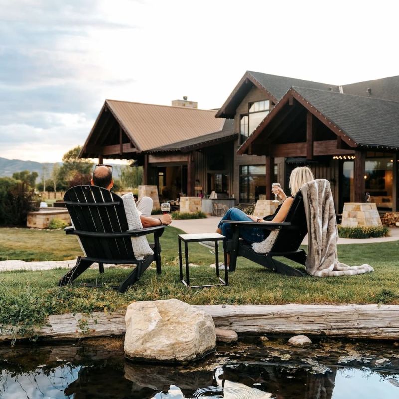 Couple in Adirondack chairs holding wine glasses beside a pond facing a timber lodge