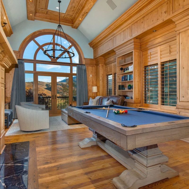Wood-paneled game room with blue-felt pool table, fireplace, and arched mountain-view doors
