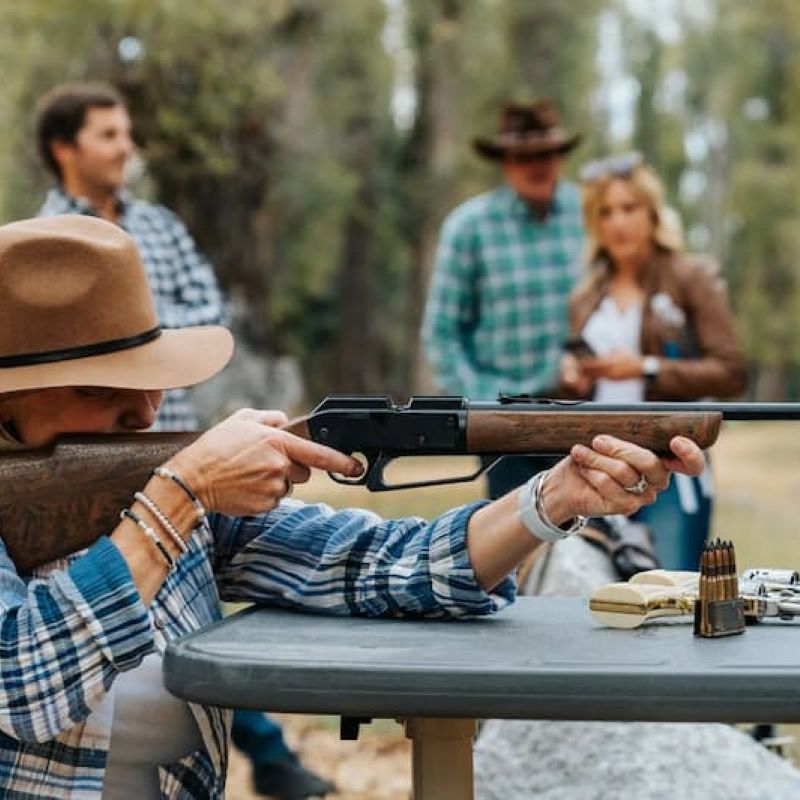 Woman shooting a lever-action rifle at an outdoor target range with onlookers