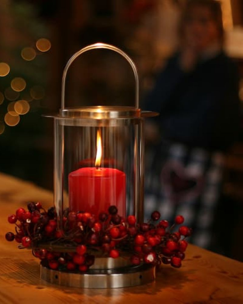 Lit red pillar candle in a lantern wreathed with red berries on a wooden ledge