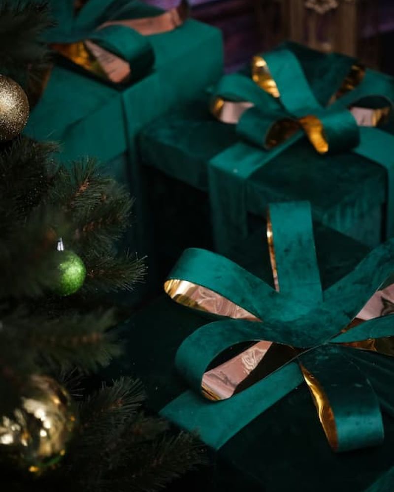 Stack of green velvet wrapped gifts with gold ribbons beside a Christmas tree