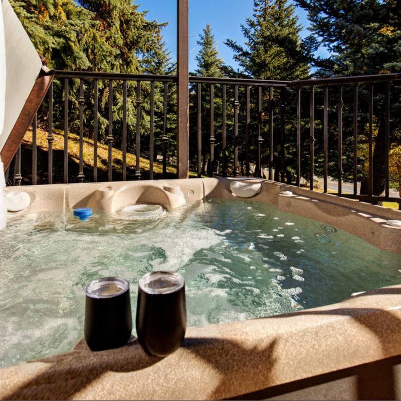 Outdoor hot tub on a deck surrounded by evergreens with two stemless wine cups on the rim